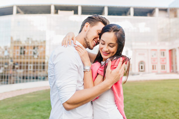 Naklejka premium Smiling girl wears trendy accessories embracing boyfriend during photoshoot on urban background. Handsome man in white shirt enjoying date with joyful brunette girl with pink sweater.