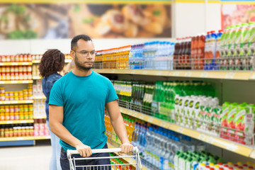 Serious African American man walking with shopping cart. Focused young man in eyeglasses making purchases in supermarket. Shopping concept