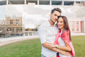 Young married couple posing together in front of modern building during joint weekend. Gorgeous dark-haired european girl huggs with boyfriend on outdoor photoshoot.