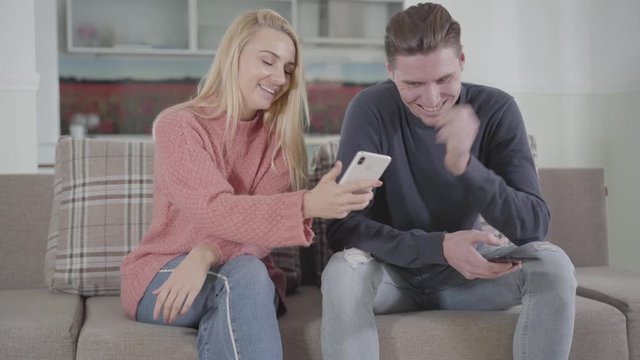 Positive Caucasian woman showing funny picture in smartphone to her boyfriend or husband. Young couple sitting on couch at home and using social media.