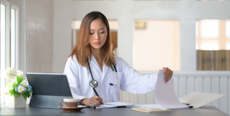 Cropped shot of female doctor working with tablet and writing on paperwork in office room