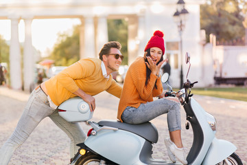 Obraz premium Shapely lady in red knitted hat sitting on scooter withfunny face expression while boyfriend pushing her. Outdoor portrait of ecstatic young people fooling around in park in warm autumn day.