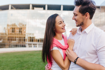 Stunning caucasian lady looking into boyfriend's eyes with tenderness and smiling in park. Lovely girl with elegant manicure holding hands with husband while posing near sparkle building.