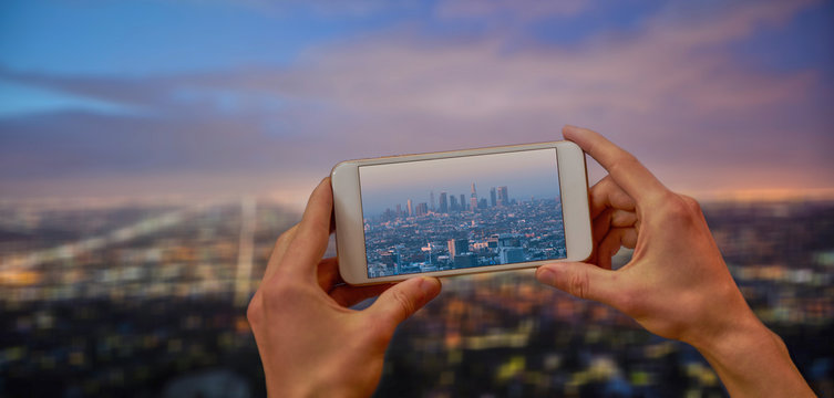 Hands holding a mobile phone and taking a picture of cityscape Los Angeles at dusk
