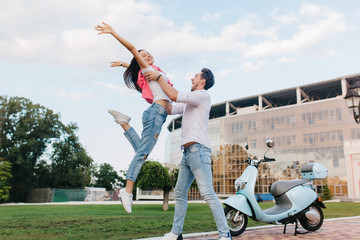 Inspired slim girl in vintage jeans jumping with hands waving while boyfriend holding her. Young people having fun on date after riding on scooter in summer day.