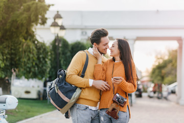 Dreamy girl in stylish orange sweater holding smartphone and gently touching boyfriend's face. Outdoor photo of smiling couple travelling around city with camera and backpack.