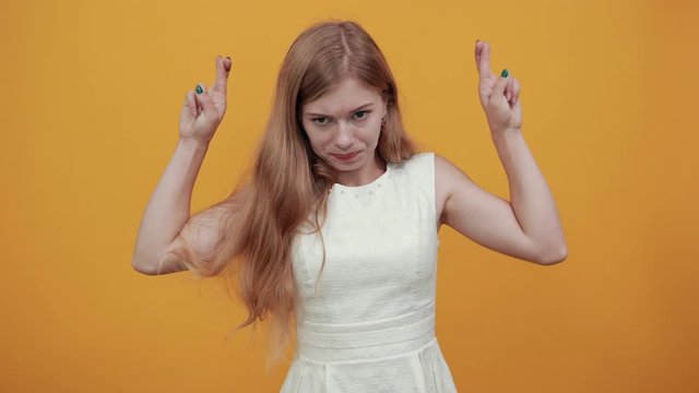 Disappointed caucasian young lady holding finers crossed, make wish, waiting for special moment over isolated orange background in blue shirt with white polka dot. Lifestyle concept