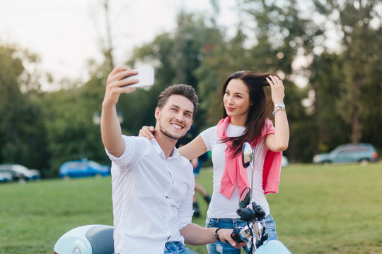 Adorable brunette girl plays with her long hair while boyfriend taking picture of her. Enthusiastic dark-haired man on scooter making selfie during meet with charming lady.