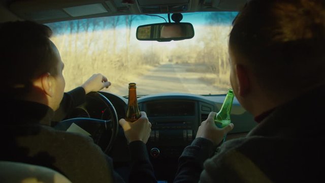Scene of two men ride in a car with bottles of alcohol, they drink alcohol while driving, chatting, laughing. The camera is behind. Drunk driver idea and concept