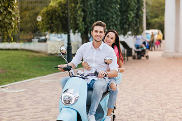 Excited man with pale skin posing on scooter with lovely dark-haired girl beside. Outdoor portrait of laughing guy in stylish white shirt drives around park with cute woman.