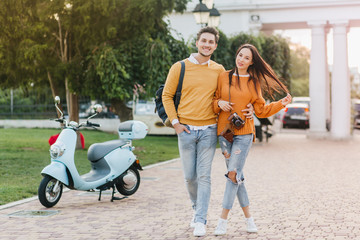 Full-length portrait of loving couple enjoying weekend together and walking in park in morning. Adorable female photographer in ripped jeans plays with her long hair while spending time with husband.