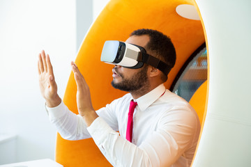 Focused businessman in VR headset. Serious young African American businessman sitting in spherical chair and using virtual reality headset. Technology concept
