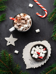Hot chocolate with marshmallows and cocoa powder, candy canes, white star and fir tree branches on dark black textured background