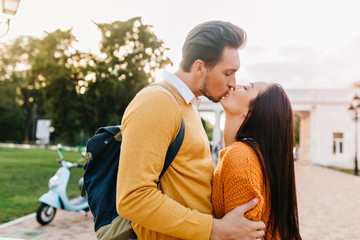 Fototapeta premium Young man with stylish haircut kissing beautiful brunette girl with eyes closed. Portrait of inspired pretty lady enjoying outdoor date with boyfriend in orange attire.