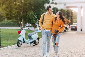 Enthusiastic girl in bright orange sweater exploring town with her boyfriend which carrying backpack. Outdoor portrait of stylish couple walking in park and talking with scooter on background.