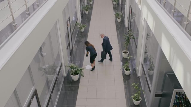 Businesswoman drops lets down documents or papers in large office hall. Colleague businessman helps her to gather and to pick them up. Awkward case in business center