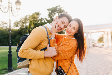 Attractive black-haired girl with elegant manicure gently touching boyfriend's face and smiling in park. Portrait of blissful young man in orange sweater with backpack enjoys free time with wife.