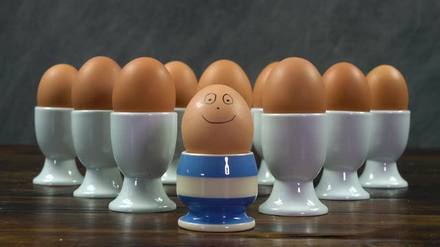 Rack focus shot of boiled egg in a striped egg cup with a smiling face in front of a group of eggs in white egg cups on a wooden table, being different, happiness concept