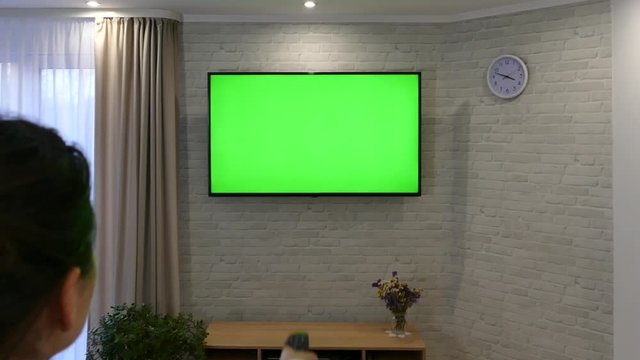 A woman is watching TV with a green screen. Switch channels with the remote control. Plasma TV hanging on the wall in the living room against a brick wall.