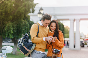 Male tourist with backpack looking at phone screen with serious face expression. Magnificent female photographer with camera standing with boyfriend on the street with green tree on background.