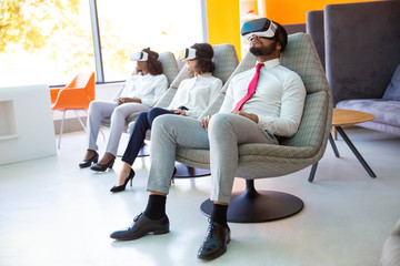 Group of multiethnic business people in VR headsets. Full length view of professional business colleagues in formal wear sitting and using virtual reality headsets in office. Technology concept