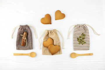 Fototapeta na wymiar Heart shaped cookies, cinnamon and spoons with cardamon on cotton bags on white wooden background. Flat lay style.