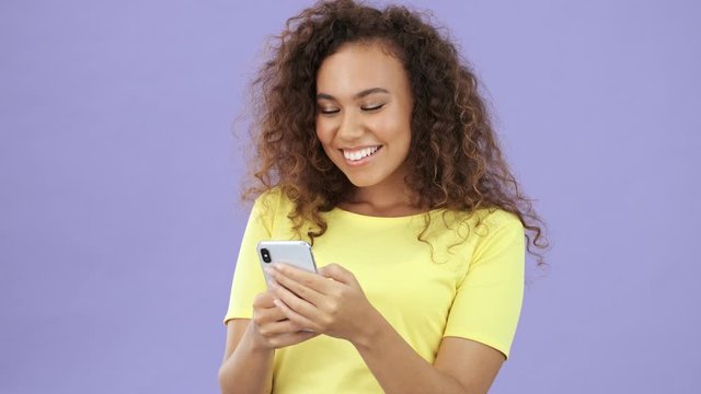 Beautiful african young woman in yellow t-shirt becoming surprised and happy because of getting some good news while using a smartphone over purple background isolated 