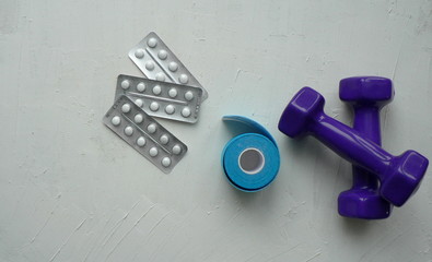 Two purple dumbbells, pills and blue kinesiology muscle tape roll on a white textured background with space for your text.