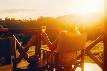 Happy woman with glass enjoys a delicious drink while sitting on the terrace during sunset.