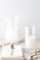 A glass and a jug of milk with metal spoon on a white napkin and pussy willow on a wooden table on white background