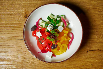 Greek salad with tomatoes, feta cheese, onions, cucumbers and bell peppers in a white bowl on a wooden background. Top view. Healthy food. Fitness nutrition and diet