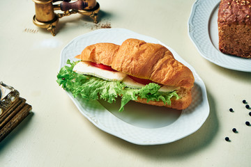 Tasty snack - croissant with chicken, cheese, tomatoes and lettuce on white plate and white wooden background. Close up view.