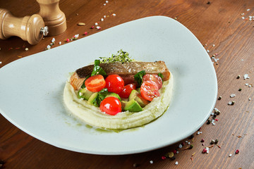 Salmon steak on a pillow of mashed potatoes with avocado and cherry tomatoes in a white plate on a wooden background. Close up view with copy space.
