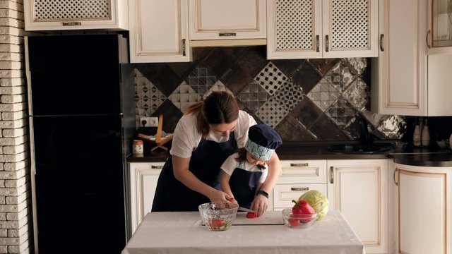 A young woman with a small daughter is preparing a vegetable salad in the kitchen, her daughter helps her mother cut a tomato. Cute three-year-old girl learns with her mother to cut vegetables.