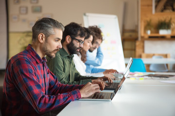 Startup team sitting at meeting table, using laptops. Business colleagues in casual working together in contemporary office space. Collaboration concept