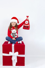 Emotional portrait of a little girl in a Santa Claus hat sitting on a cube with a bag of gifts in her hands, on a white background. The concept of a happy childhood. Blank space for text. Mock up
