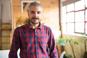 Friendly entrepreneur posing in modern office. Man with moustache in casual shirt and glasses standing in contemporary office space. Startup leader concept