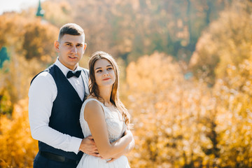 Portrait of cute newlyweds standing on a hill against the background of autumn forest.