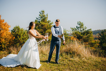 Pretty newlyweds are standing on a hill on a background of autumn forest. The groom opens a bottle of champagne