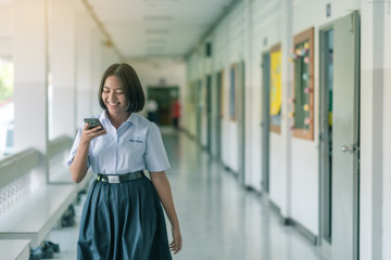 A smiling Asian female high school student in white uniform is walking and enjoying social media on her smartphone in the school.