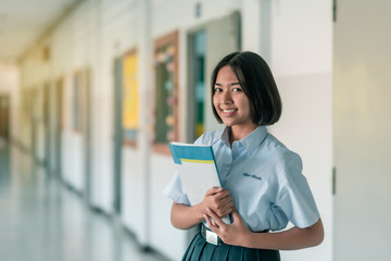 The smiling Asian female high school students in white uniform is standing and holding a book on the corridor in the school building