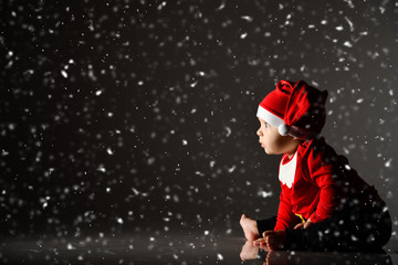 Fototapeta na wymiar Infant baby boy toddler in red christmas cap and new year costume is sitting on ice under the snow staring at light ray