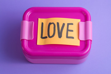 Pink lunch box with orange sticker on purple background. Love message sticker. Lunch at school from a loving wife or mother with a note. Bright colours and care concept. Idea food from home packaging.