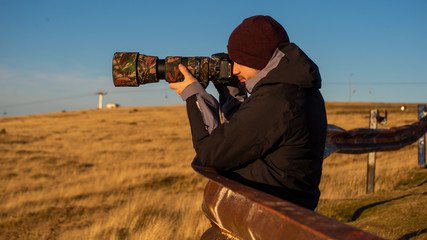 Man photographer holding camera with long lens and taking pictures in outdoors during sunny autumn day.