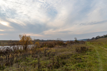 Fototapeta na wymiar Autumn landscape. A vague spot of the setting sun in the sky with wavy stripes of clouds. Valley of the river, overgrown with withering grass and bushes. A curved dirt road runs along the shore