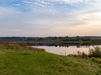 Calm landscape at eve dusk. Green bank with a place for relaxation and a picnic with beautiful view of the river, distant forest. The still mirror of the water reflects trees and sunset in cloudy sky