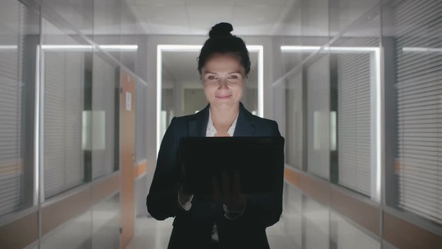 Young happy attractive caucasian businesswoman formal suit walking through office hall corridor in slow motion holding laptop computer looking for documents contract details. Lady smiling