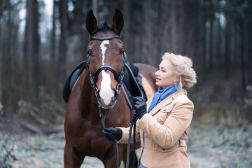 Fototapeta premium A 50 year old woman rides a horse. Walking with a horse. He is engaged in hippotherapy. A woman is wearing a trendy derby costume.