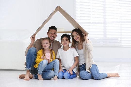 Happy Family Sitting Under Cardboard Roof At Home. Insurance Concept