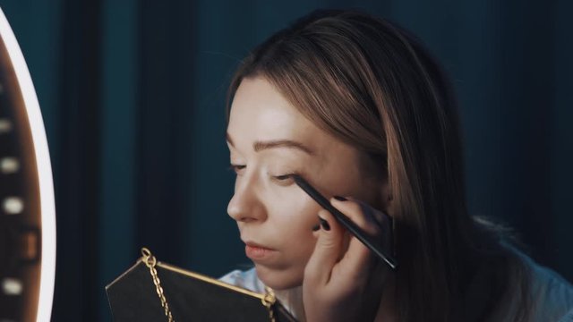 Good-looking woman beauty blogger in white shirt holds mirror and demonstrates usage of eyeliner on camera, sitting in front of ring light, filming tutorial video blog. Shot on 4K RED camera.